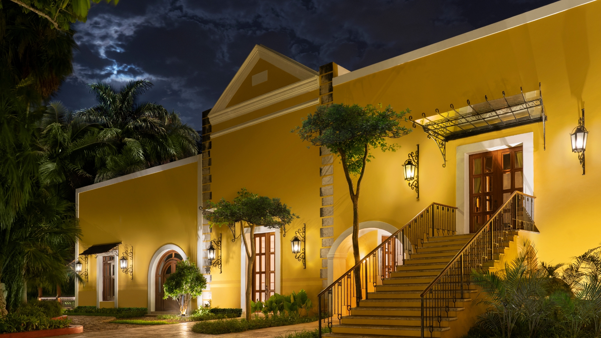 Yellow building illuminated at night, with stairs leading up, surrounded by lush trees at Hacienda Xcanatun, Angsana Heritage Collection.